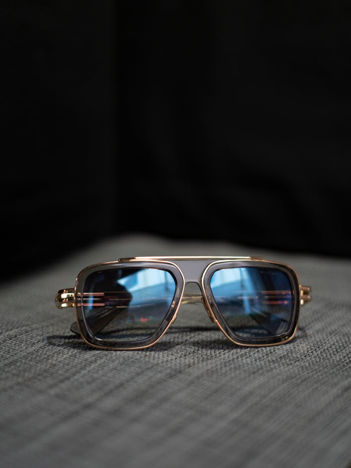 gallery-03 Elegant aviator sunglasses with blue lenses and gold frame on a textured grey surface.